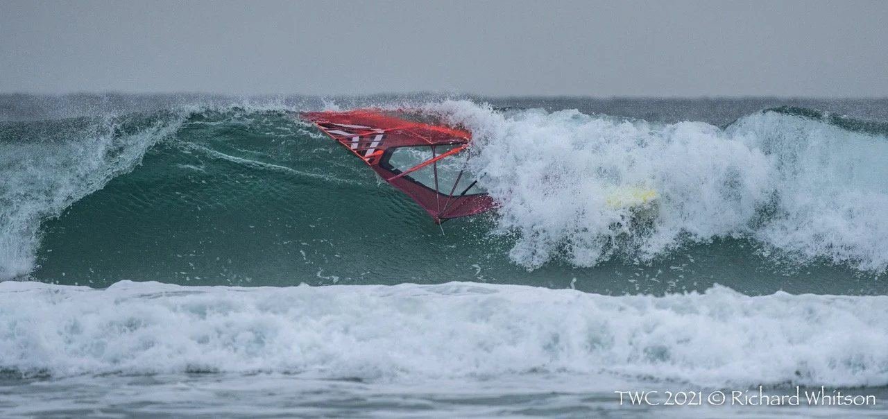 A windsurfer with a red sail drops into a hollow green wave