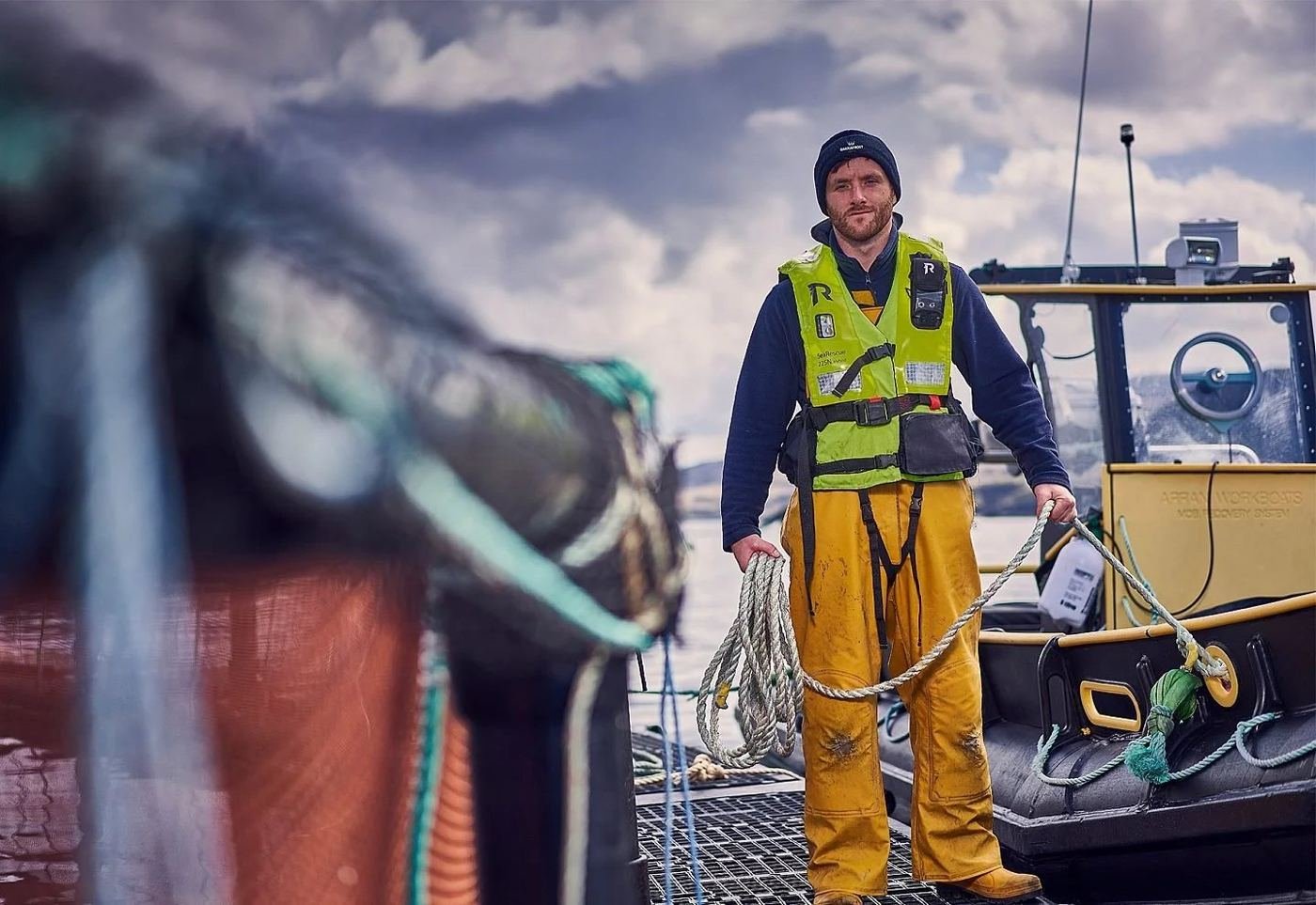 A Bakkafrost fish farmer in yellow waterproofs beside a workboat